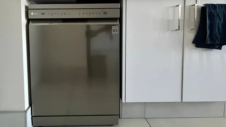 A stainless steel dishwasher is integrated into a modern kitchen layout, surrounded by white cabinets. A blue towel hangs on a cabinet handle nearby. The dishwasher features digital controls on the front panel. Dishwasher Plumbing