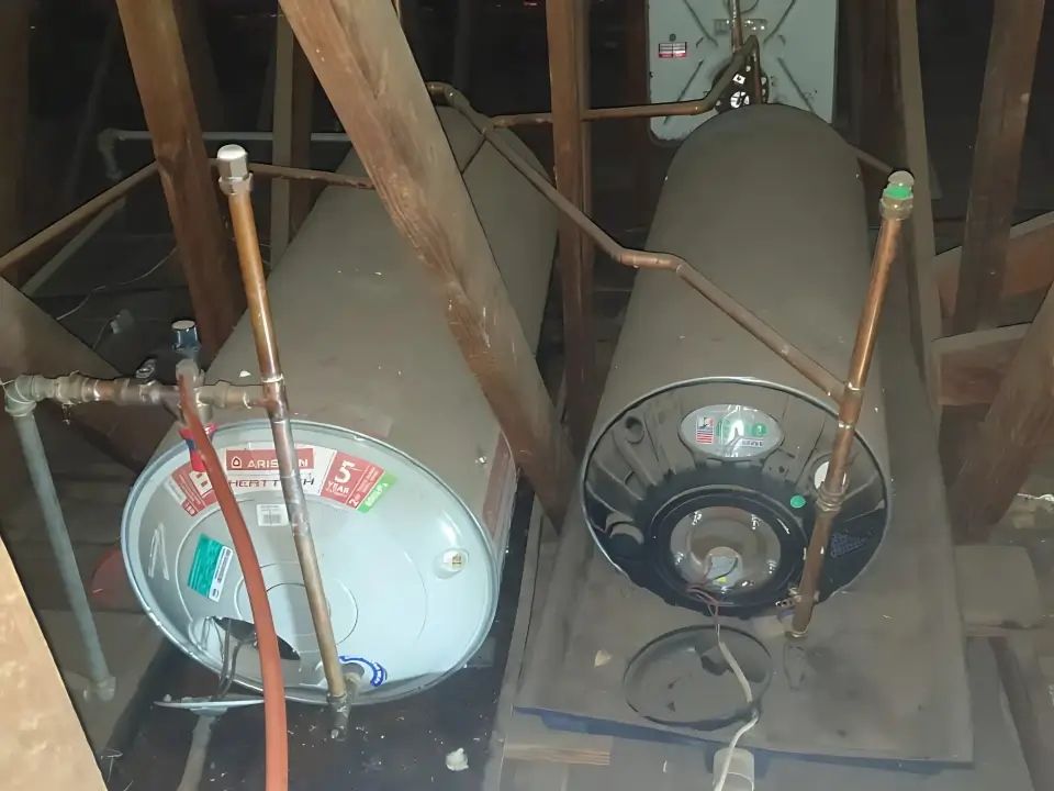 Two cylindrical water heaters are positioned horizontally in an attic. They are surrounded by wooden beams and piping, with each heater having visible labels and connections. The space appears to be dimly lit. Installed Water Heater also Known as Geyser In South Africa