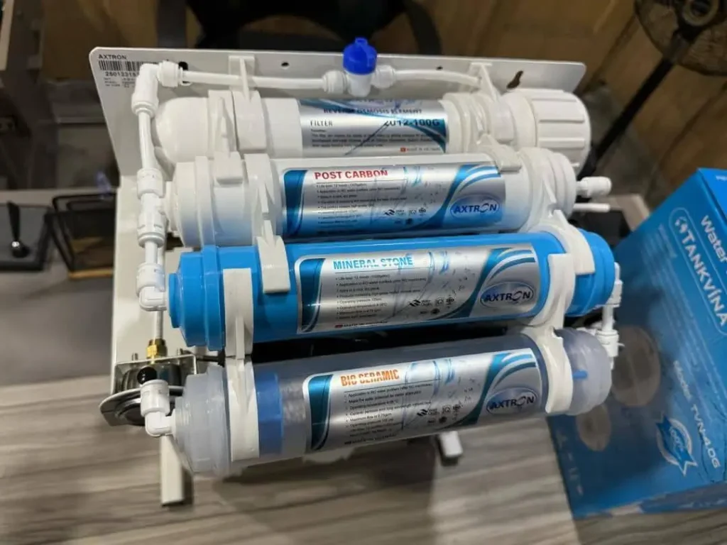 A close-up of a multi-stage water filtration system featuring several cylindrical filter cartridges labeled Post Carbon, Mineral Stone, and Bio Ceramic. The setup is mounted on a white frame. Home Water Filtration System