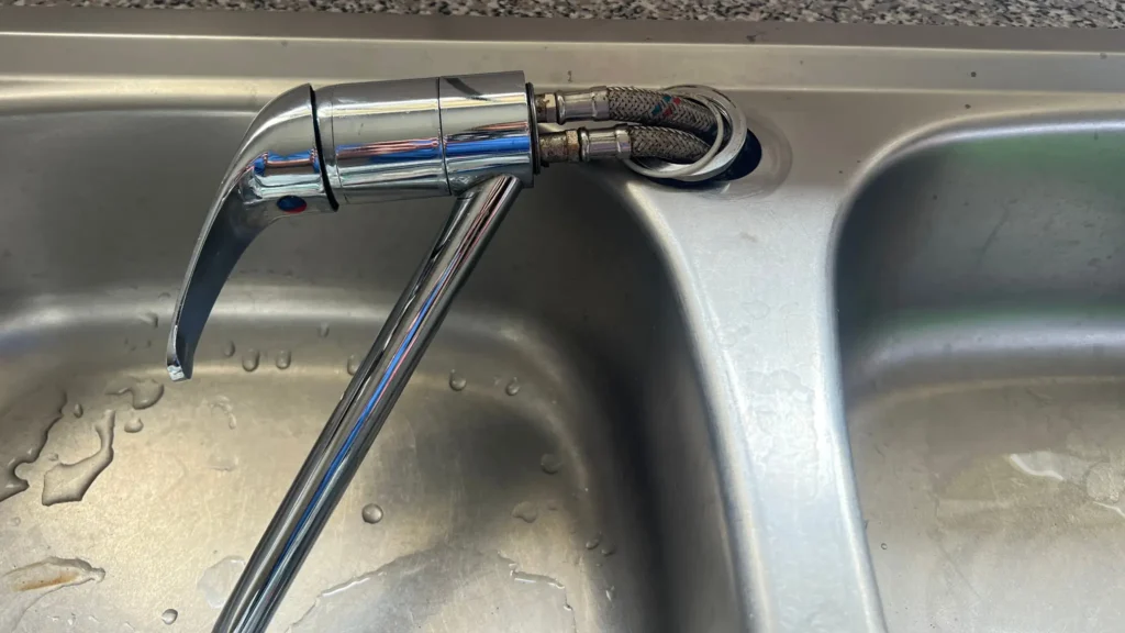 A damaged kitchen sink faucet with the fixture detached and the hoses exposed, resting on a dual-basin stainless steel sink. Water droplets are visible on the left basin. Kitchen Plumbing