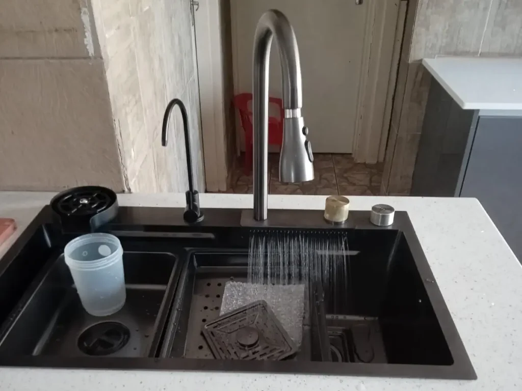 A kitchen sink with a black basin features a modern, curved faucet. Water is flowing from the faucet into the sink. Inside are a clear plastic cup and a black strainer. The countertop is light-colored, contrasting with the dark sink. Kitchen Plumbing Services