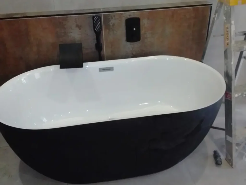 A modern bathroom with a sleek, freestanding black and white bathtub. The wall behind features a panel with a minimalist black faucet and handheld shower. A ladder is partially visible to the right. Bathroom Plumbing Services
