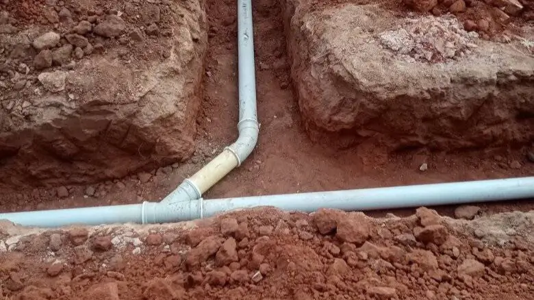 A set of white PVC pipes are laid in dirt trenches, forming a T-junction. The surrounding soil is reddish-brown, with uneven surfaces and exposed rock edges. Pipe Repairs & Replacements.