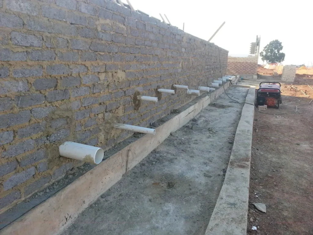 A partially constructed brick wall with several white PVC pipes protruding horizontally. The pipes are evenly spaced along the wall. In the background, construction materials and a small generator are visible on the dirt ground. Pipe Repairs & Replacements.