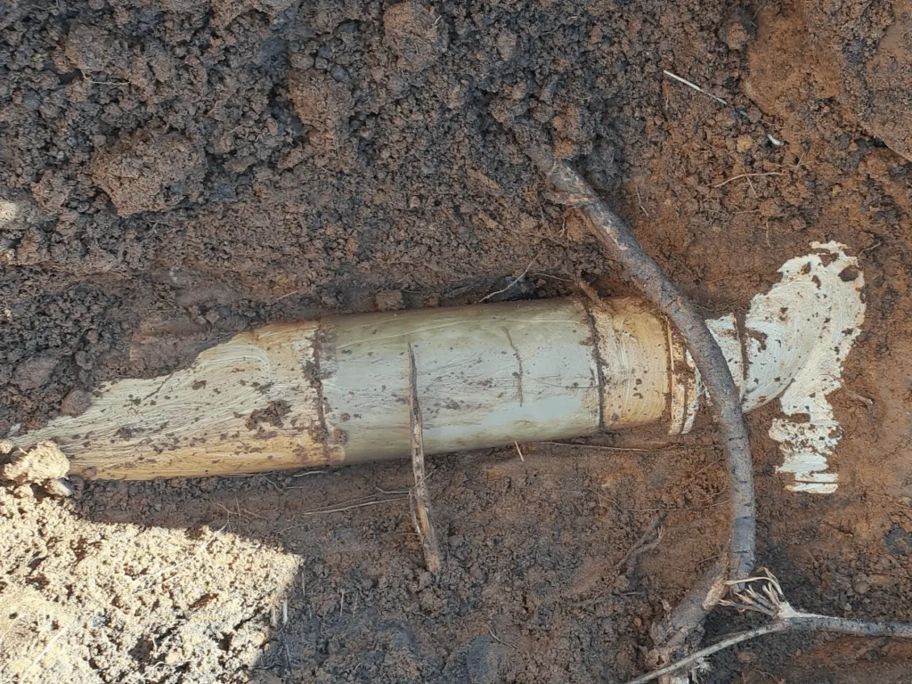 A partially unearthed old artillery shell lies in the dirt, surrounded by dark soil and a small stick. The shell is corroded and weathered, blending with the earth around it. Sewer Line Leak Investigation