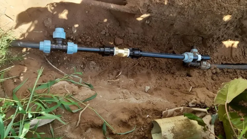 A section of PVC piping with connectors is installed in a dirt trench. The surrounding area has soil and some green grass. The assembly includes various connectors and a brass fitting. Nearby are leaf litter and a piece of paper or plastic. Sewer Line Leak Repair