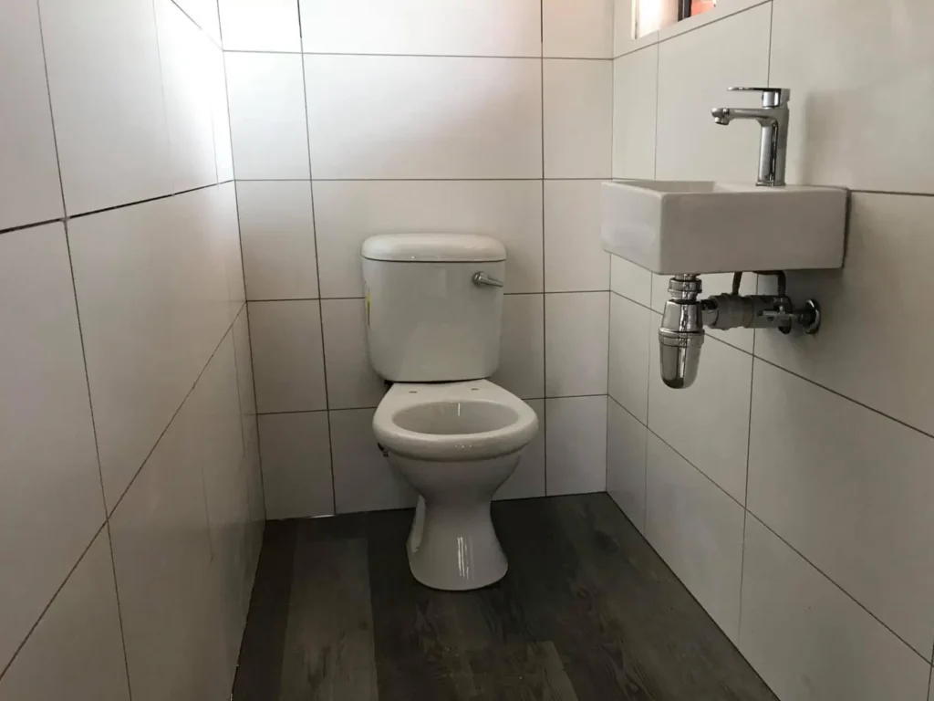 A small bathroom with white tiled walls and wood-patterned flooring features a toilet and a compact white sink with a metallic faucet. The space appears clean and minimalist. Toilet Plumbing - New Toilet Installation