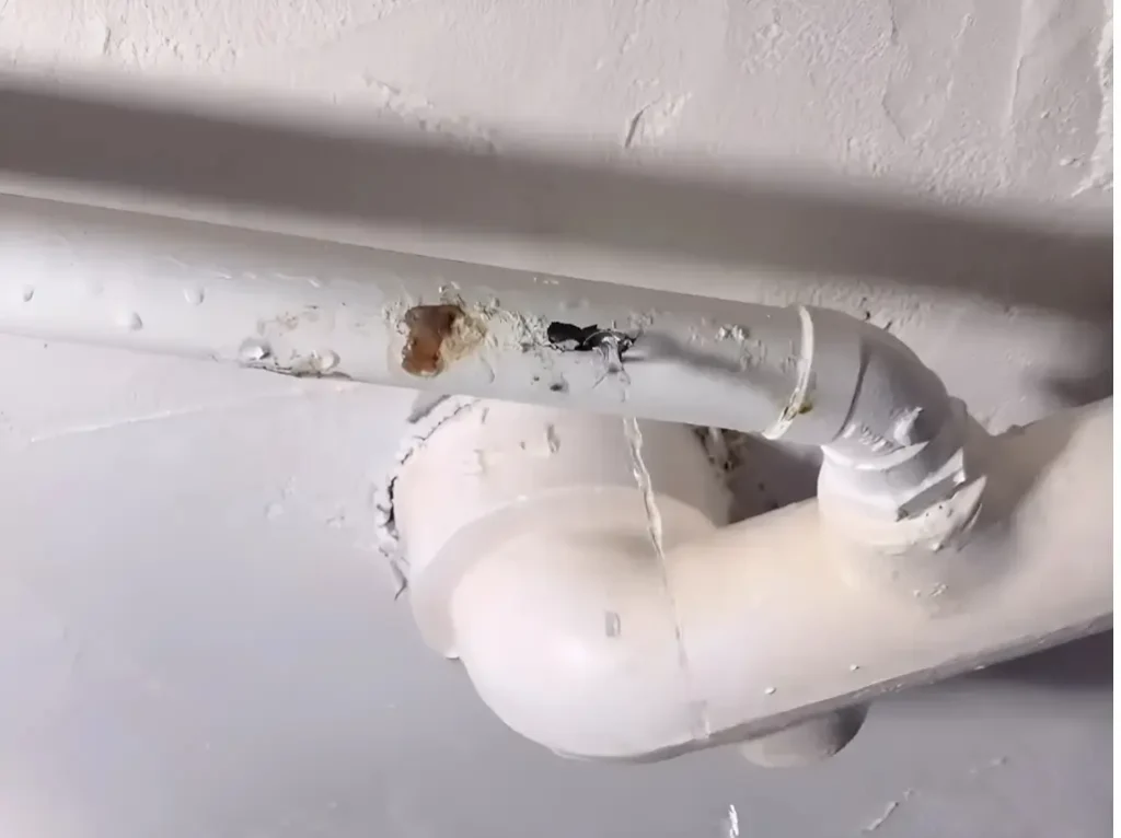 A white PVC pipe with a bend is leaking water from a hole and a crack, forming a steady stream. The ceiling around the pipe shows signs of moisture damage. This requires Emergency Plumbing Services.