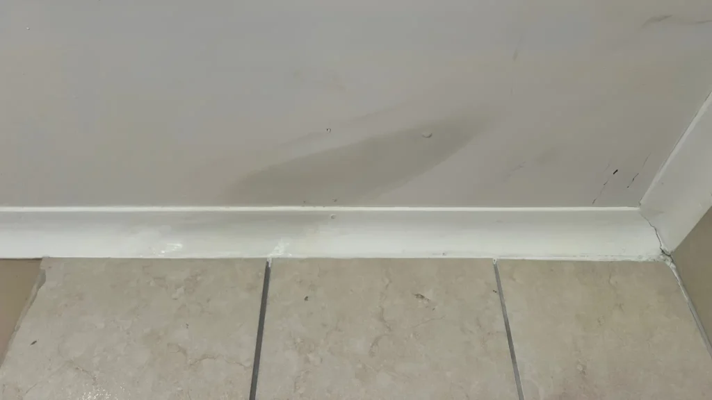 leak on the ceiling. A section of a wall with visible water damage and stains near the baseboard. The flooring consists of beige tiles with grout lines. The wall and baseboard show signs of moisture and discoloration. Leak Detection