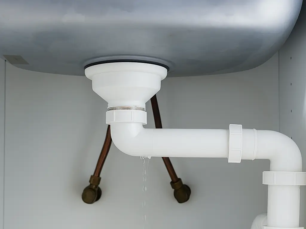 A close-up of the underside of a sink shows white plastic plumbing with a slow drip of water coming from a joint. Two copper pipes are visible in the background against a white cabinet interior. Leak detection image, showing a leak under the sink