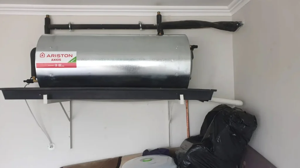 A silver heater tank labeled ARISTON AXIOS is mounted on a wall shelf in a corner. Pipes are connected to it, and nearby are bags and part of a circular item. The wall is plain with white trim. water Heater Installed